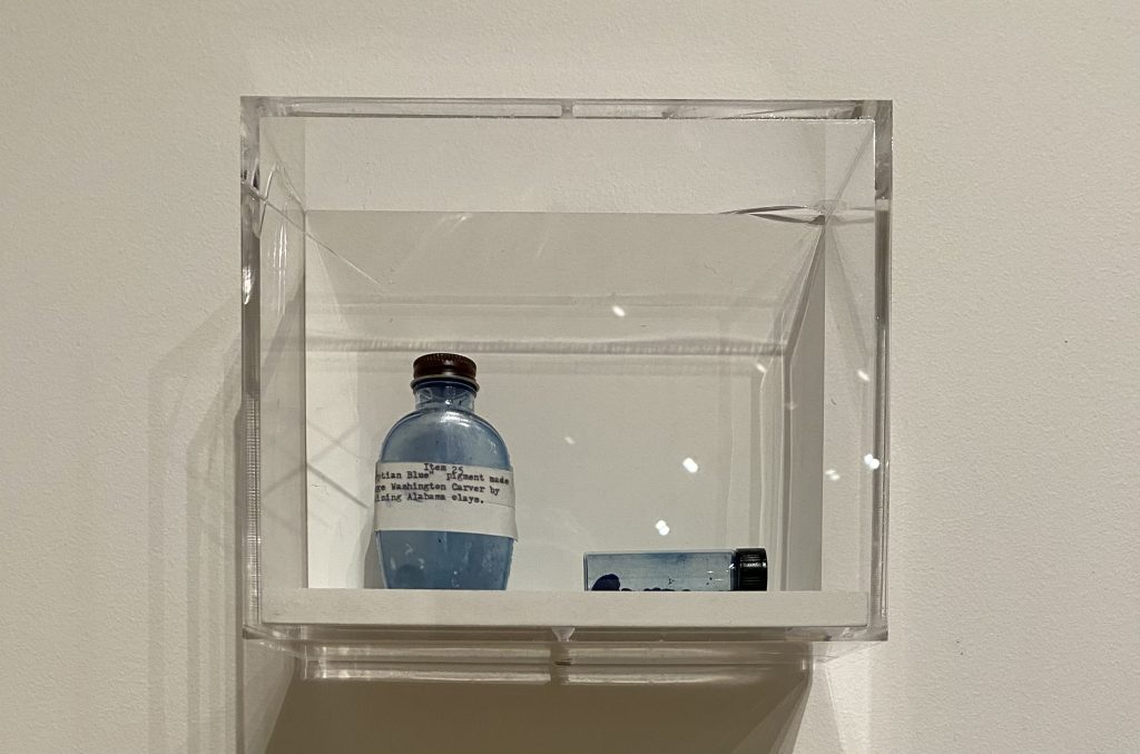 A small glass bottle filled with bright blue pigment is displayed inside a clear acrylic case mounted on a gallery wall.