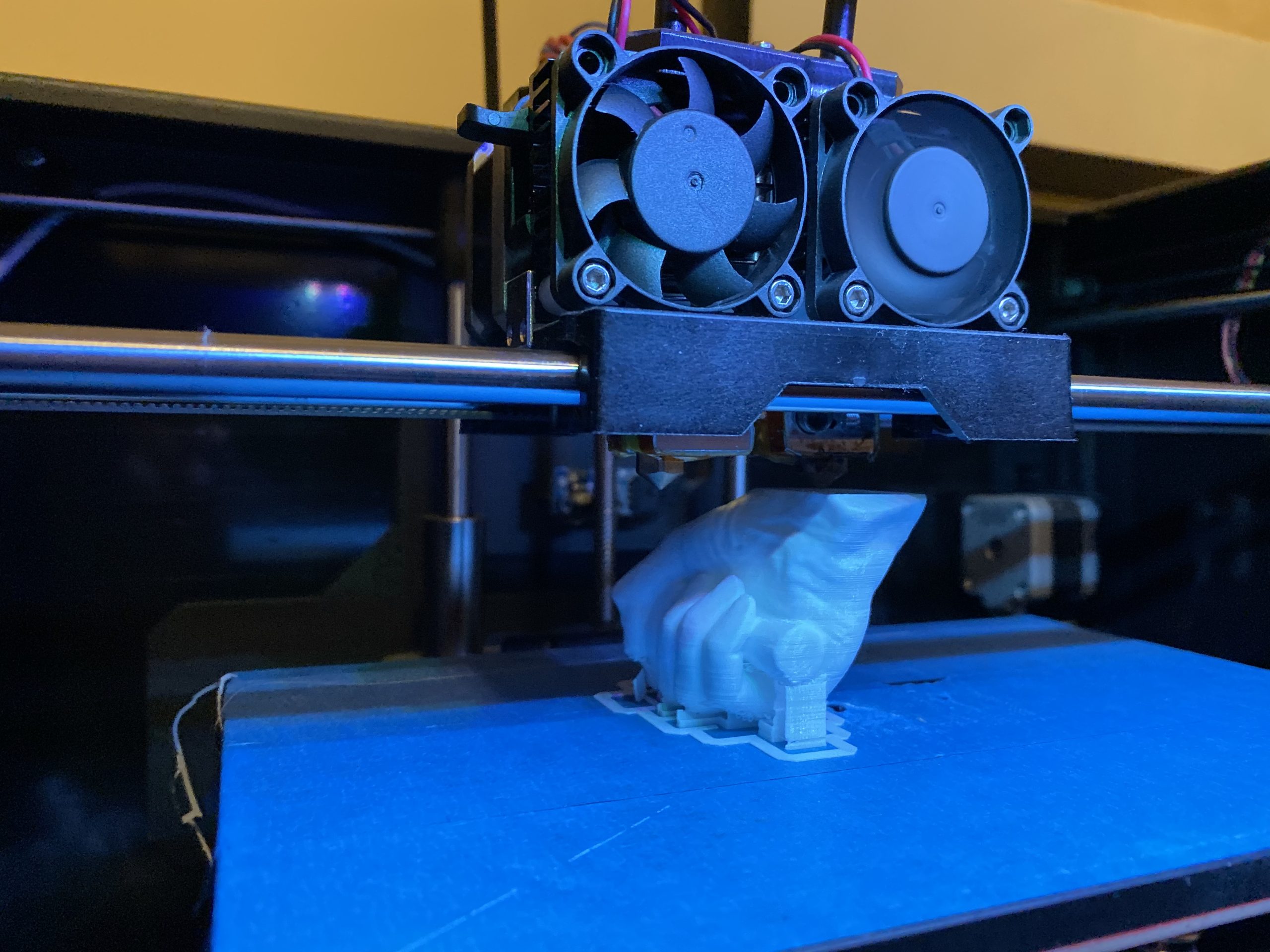 A 3D printer mid-process, printing a translucent blue model of a human hand gripping a cylindrical object. The extruder head and cooling fans are visible above the partially completed print.