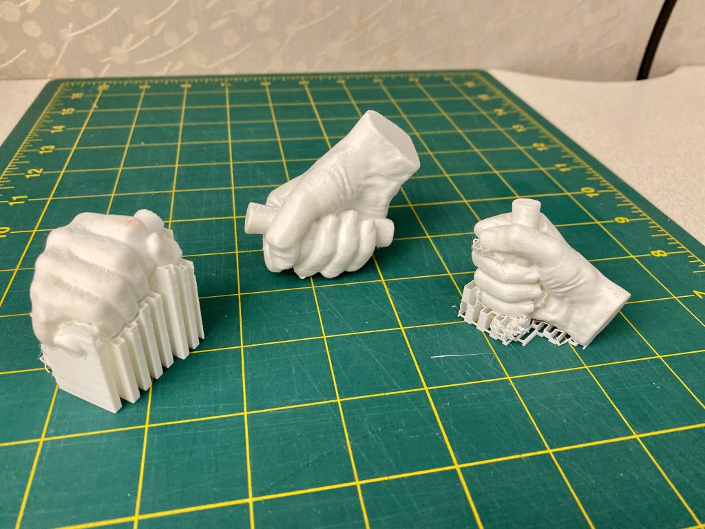 Three 3D-printed replicas of a human hand gripping a cylindrical object, displayed on a green cutting mat. Two prints still have support structures attached, while the center model is fully cleaned and finished.