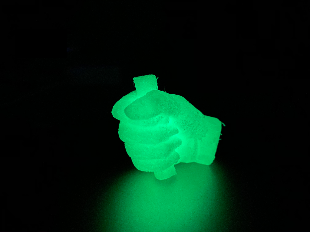 A 3D-printed model of a human hand gripping a cylindrical object, glowing green in the dark against a completely black background.