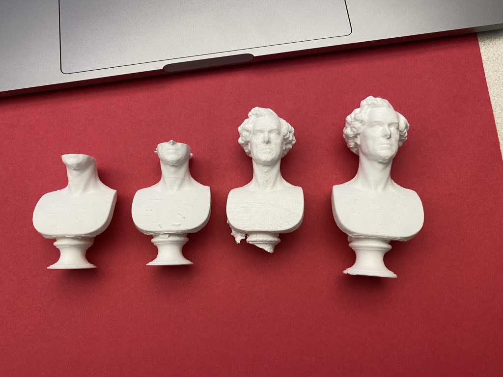 Four 3D-printed miniature busts in progressive states of completion or failure, arranged left to right on a red background beneath a laptop. The first two prints lack heads, the third is partially intact with surface issues, and the fourth is a clean, fully printed bust.