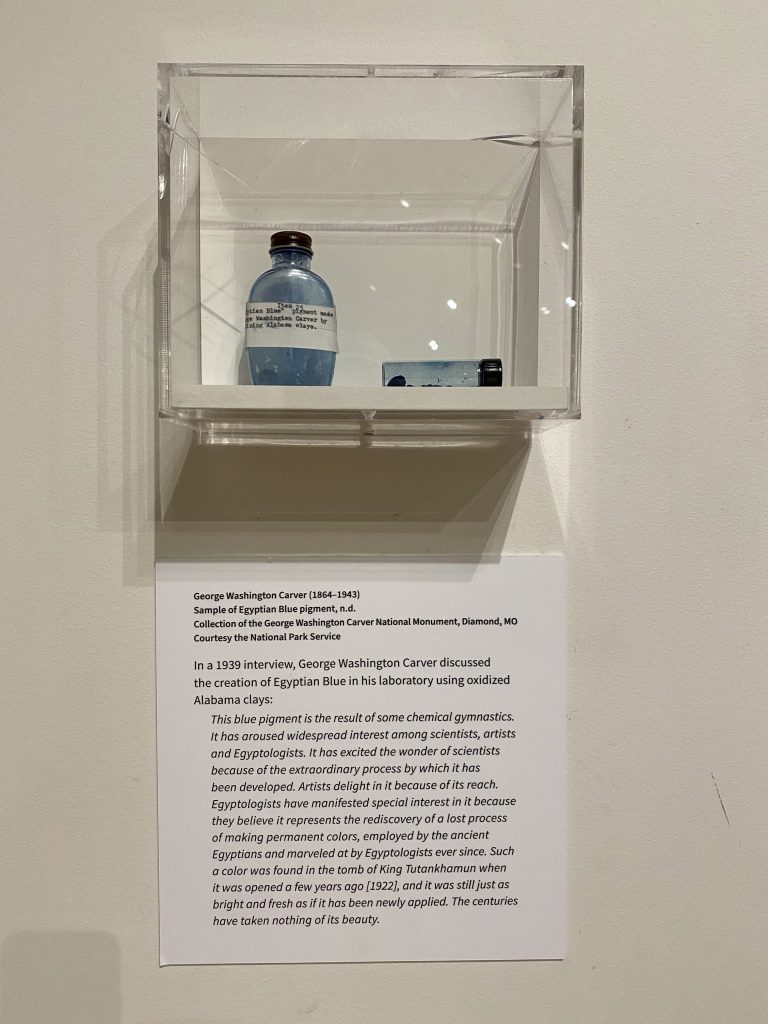 A small glass bottle filled with bright blue pigment is displayed inside a clear acrylic case mounted on a gallery wall. Below it, a wall label explains the pigment’s creation using oxidized Alabama clay, highlighting its connection to ancient Egyptian color-making techniques and quoting a 1939 interview about its scientific and artistic significance.