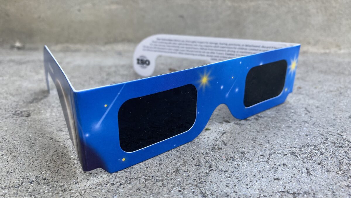 A pair of ISO-certified solar eclipse glasses with a blue starry design, resting on a concrete surface. The dark protective lenses and printed certification markings are visible.